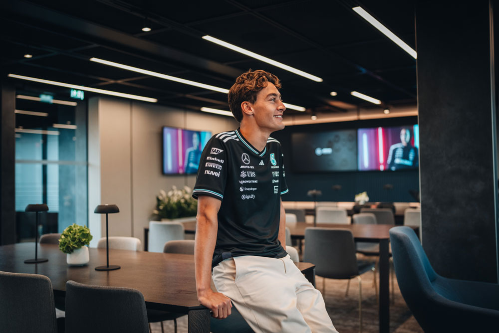 George Russell, piloto del equipo Mercedes-AMG PETRONAS Formula One Team, está sentado de manera relajada sobre una mesa en una zona moderna y elegante del equipo, ya sea una oficina o un área de hospitalidad. Sonríe y lleva puesta la camisa oficial del equipo con los logotipos de los patrocinadores visibles, como Einhell, IWC y CrowdStrike. En las pantallas del fondo aparece su imagen.