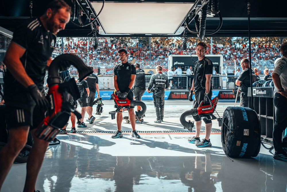 Los mecánicos del equipo Mercedes-AMG PETRONAS Formula One Team se preparan en el garaje con sopladoras inalámbricas Einhell antes de una parada en boxes. Al fondo, las tribunas están repletas de aficionados.