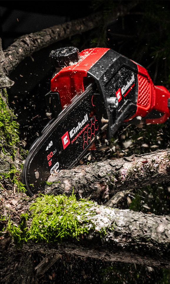Una motosierra inalámbrica roja de Einhell corta una rama mientras el serrín vuela por el aire.